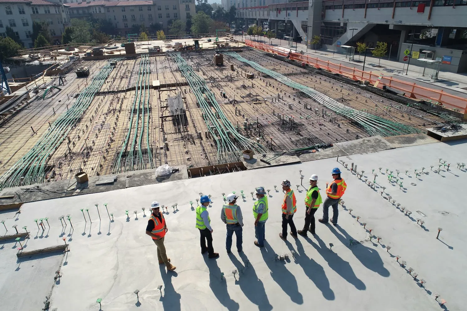 Seven people in high-vis vests and hardhats, having a discussion while looking out over a large construction site in a metropolitan area
