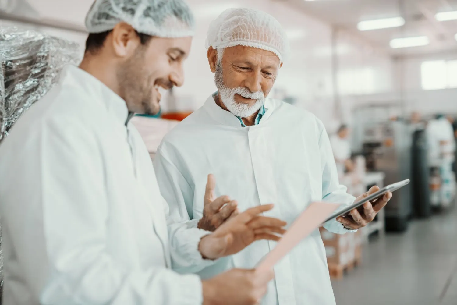 Two food production workers in a commercial kitchen, wearing hair-nets white coats while reading a tablet screen