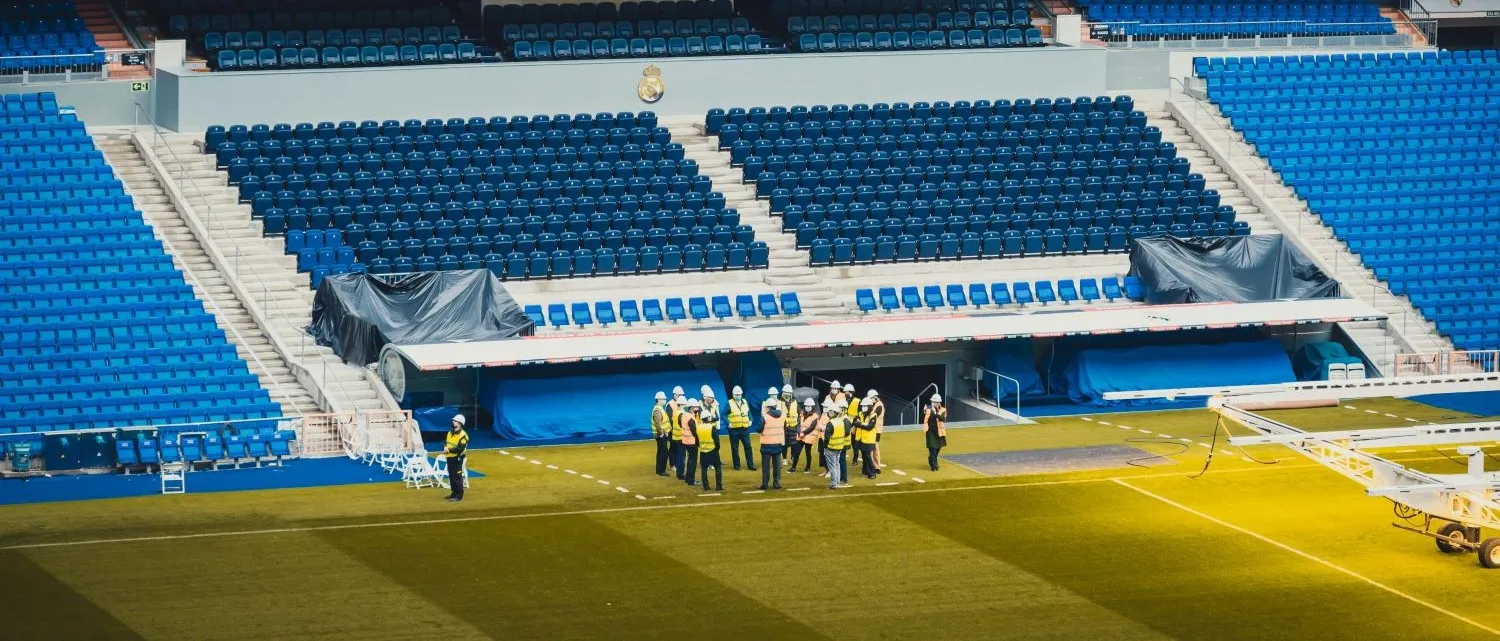 A group of employees in hi-vis standing inside a large empty sports stadium stadium, listening to a safety briefing