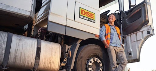 A DSE Transport worker in an orange safety vest. He is leaning against a white truck cab with the driver's door open. The image represents DSE's improvements to worker induction and onboarding.
